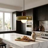 A modern kitchen scene with a hanging pendant light above a marble island. The light has a translucent olive-green glass shade, a dark brown top cap, and a glowing white bulb.