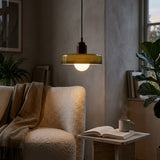 A modern pendant light with a translucent olive-green glass shade and a dark reddish-brown metal top, glowing warmly in a cozy living room setting next to a textured beige armchair and a small side table with an open book and mug.