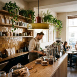 A warm, cozy cafe interior with a barista preparing coffee behind a wooden counter. The space is decorated with numerous green trailing plants on wooden shelves and the counter, a rustic brown tiled backsplash, and a prominent amber-green dome pendant light hanging over the workspace.