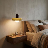 A modern pendant light with a translucent olive-green shade and a dark brown cylindrical neck hangs above a floating wooden nightstand in a bedroom setting. The room features a textured plaster wall, a bed with neutral beige linens, and warm ambient lighting.