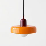A modern pendant light with a glossy orange ring-shaped shade, a dark red central collar, a black hanging cord, and a white globe bulb below, shown against a plain white background.