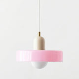 A modern pendant light with a wide, flared pink shade, a cream-colored cylindrical body, a white bulb holder, and a brass-colored connector on a white cord, set against a plain white background.