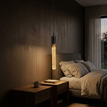 A modern cylindrical pendant light with a black top and bottom and a glowing textured glass center, hanging in a dark, contemporary bedroom next to a wooden nightstand and bed.