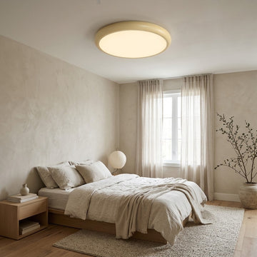 A modern flush-mount ceiling light with a warm cream-colored frame and glowing warm white light, installed in a minimalist beige bedroom with a wooden bed, textured walls, and sheer curtains.