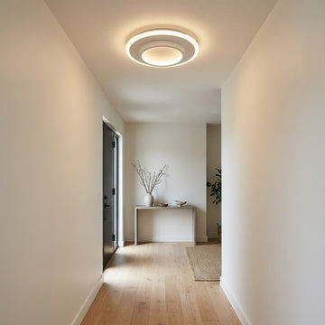 A modern circular LED ceiling light with a warm white glow, installed on a white ceiling in a minimalist hallway with light wood floors and beige walls.