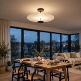 A modern circular ceiling light fixture with a frosted white glass shade and dark metal accents, mounted on the ceiling of a contemporary dining room with large windows overlooking a city view at dusk.