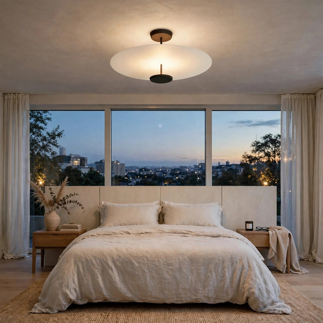A modern ceiling light fixture with a wide, shallow white glass shade and a black central diffuser, mounted on a textured ceiling in a beautifully styled bedroom with a bed, nightstands, and large windows overlooking a city skyline at dusk.