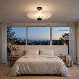 A modern ceiling light fixture with a wide, shallow white glass shade and a black central diffuser, mounted on a textured ceiling in a beautifully styled bedroom with a bed, nightstands, and large windows overlooking a city skyline at dusk.