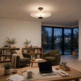 A modern ceiling light fixture with a circular white frosted shade and dark metal hardware, mounted on the ceiling of a cozy, warmly lit living room with green armchairs, wooden bookshelves, and a wooden desk in the foreground.