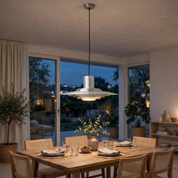 A modern silver pendant light with a tiered, saucer-like design hangs over a wooden dining table set for six in a stylishly decorated dining room with large windows and plants.