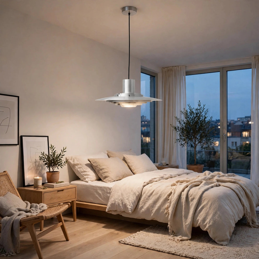 A modern silver pendant light with a wide, flared disc-shaped shade hangs over a neatly made bed in a softly lit, minimalist bedroom with beige bedding, wooden furniture, and large windows.