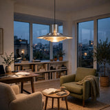 A modern silver tiered pendant light hangs in a cozy, well-lit living room with a green armchair, a wooden desk, and large windows overlooking a city skyline at dusk.