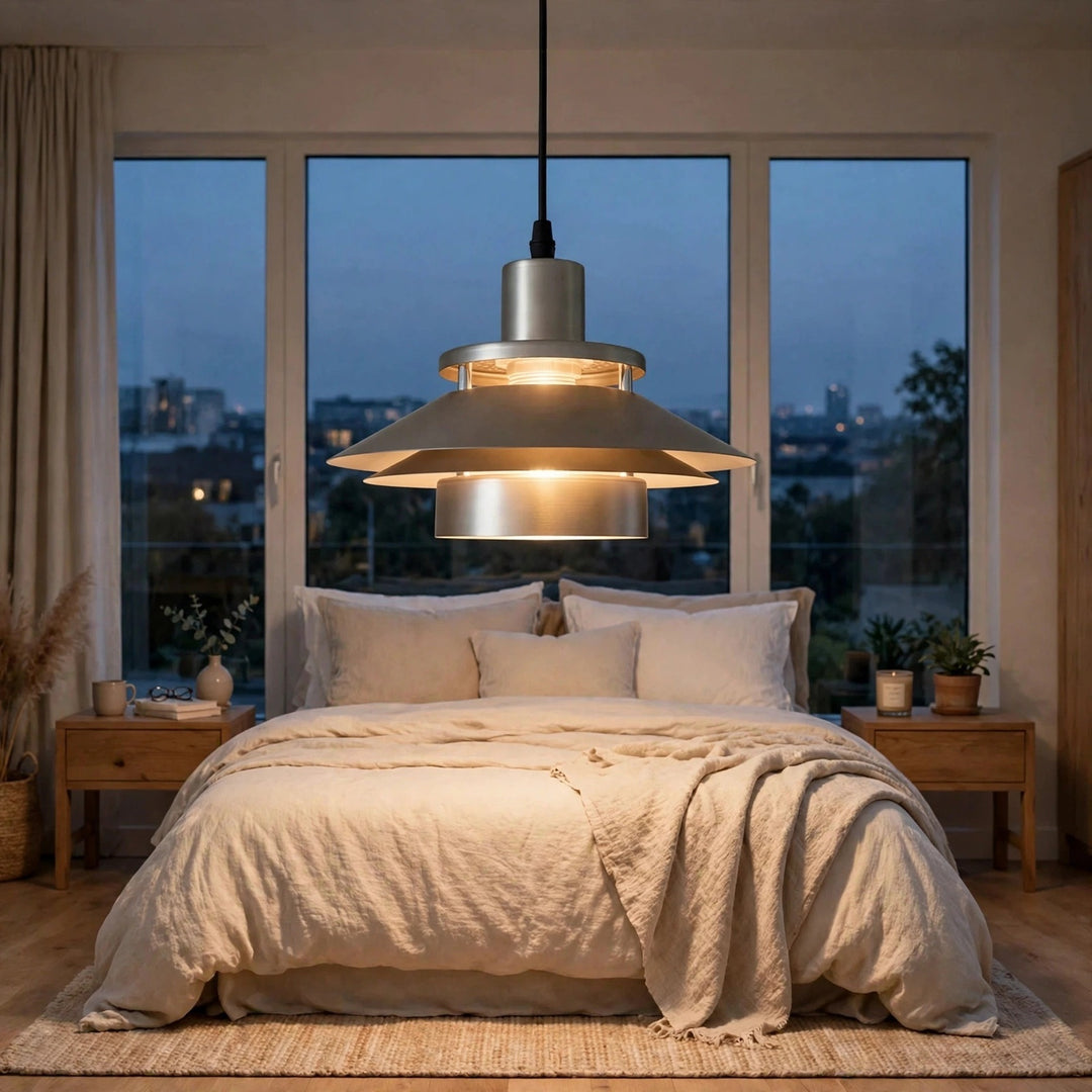 A modern multi-tiered pendant light with a brushed silver metal finish and warm glowing bulbs, hanging in a beautifully styled bedroom with a bed of beige linens and large windows showing a city view at dusk.