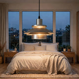 A modern multi-tiered pendant light with a brushed silver metal finish and warm glowing bulbs, hanging in a beautifully styled bedroom with a bed of beige linens and large windows showing a city view at dusk.
