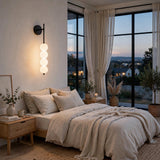 A modern wall-mounted linear sconce with four stacked spherical glass shades glowing warm white, mounted on a beige wall in a styled bedroom with a bed, nightstand, and large windows overlooking a city at dusk.