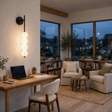 Black iron wall sconce with four glowing milky white globes mounted above a wooden desk in a living room at dusk.