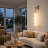 Gold wall sconce with four glowing milky white globes mounted on a beige wall above a cream living room sofa.