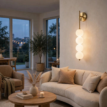 Gold wall sconce with four glowing milky white globes mounted on a beige wall above a cream living room sofa.