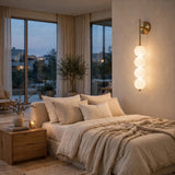 Gold wall sconce with four glowing milky white globes on a beige bedroom wall beside a bed with cream bedding.