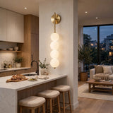 A modern vertical wall sconce with four stacked frosted glass spheres and a brushed brass top mount, shown lit in a contemporary kitchen and living room interior.
