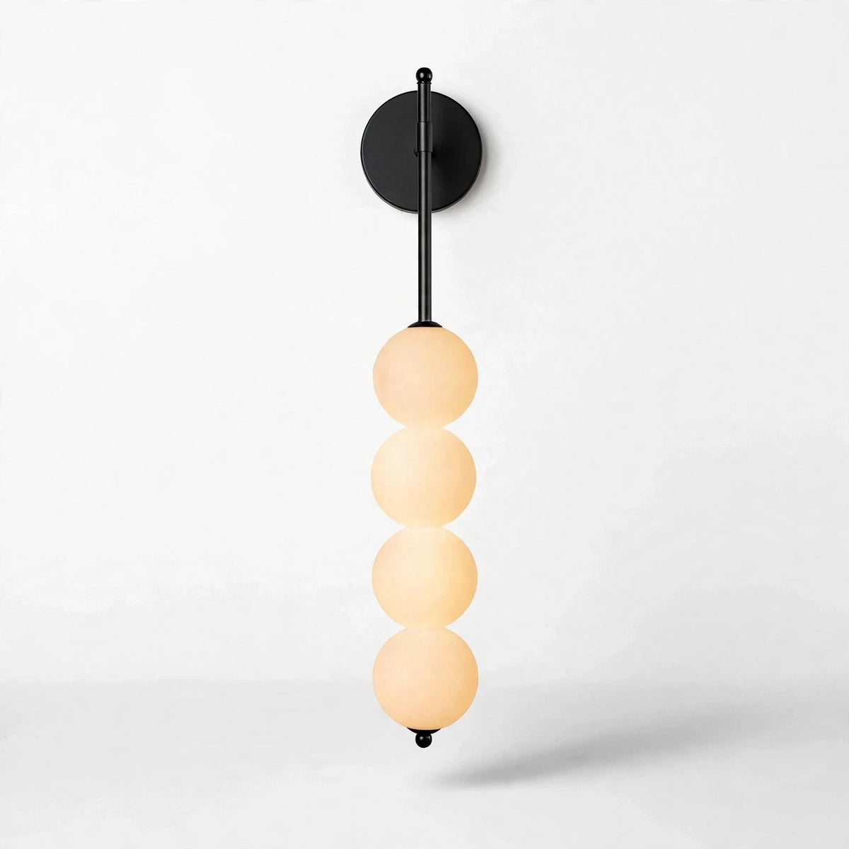 Black iron wall sconce with a round backplate and four vertically stacked milky white globes glowing warmly against a white background.