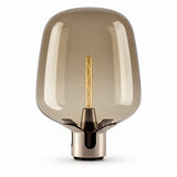 A modern table lamp with a large, rounded translucent smoke-tinted glass shade and a metallic brushed brass base, featuring a visible warm-toned filament bulb inside, set against a plain white background.
