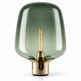 A modern table lamp with a translucent smoky green glass shade and a brushed brass metal base, featuring a visible warm-toned filament bulb inside, set against a plain white background.