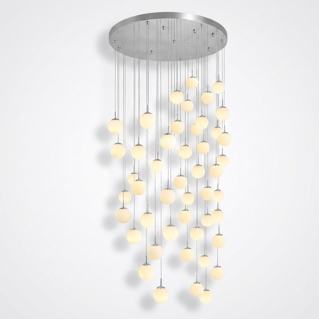 Modern multi-light chandelier with a circular brushed silver ceiling canopy and numerous hanging glass globes emitting a warm yellow glow, shown against a plain light gray wall.