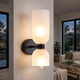 A modern two-light wall sconce mounted on a textured beige wall in a living room setting. The fixture features two frosted glass shades, one cylindrical and one bell-shaped, with a matte black metal base and arm.