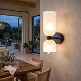 A modern wall-mounted double-light sconce with two frosted cylindrical glass shades and a dark metal base, installed on a textured beige wall in a warmly lit dining room interior.
