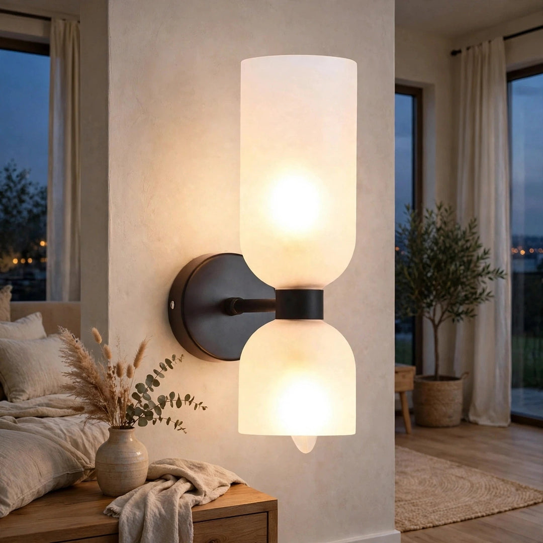 A modern two-light wall sconce with frosted cylindrical and dome-shaped glass shades, mounted on a dark metal wall plate in a warmly lit bedroom setting.