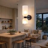 A modern two-light wall sconce with frosted cylindrical shades and a dark metal mount, illuminated in a warm glow, mounted on a wall in a contemporary, neutral-toned living and kitchen space.