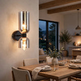 A modern wall-mounted sconce with two clear glass shades and warm exposed bulbs, mounted on a beige wall in a styled dining room with a wooden table and chairs.
