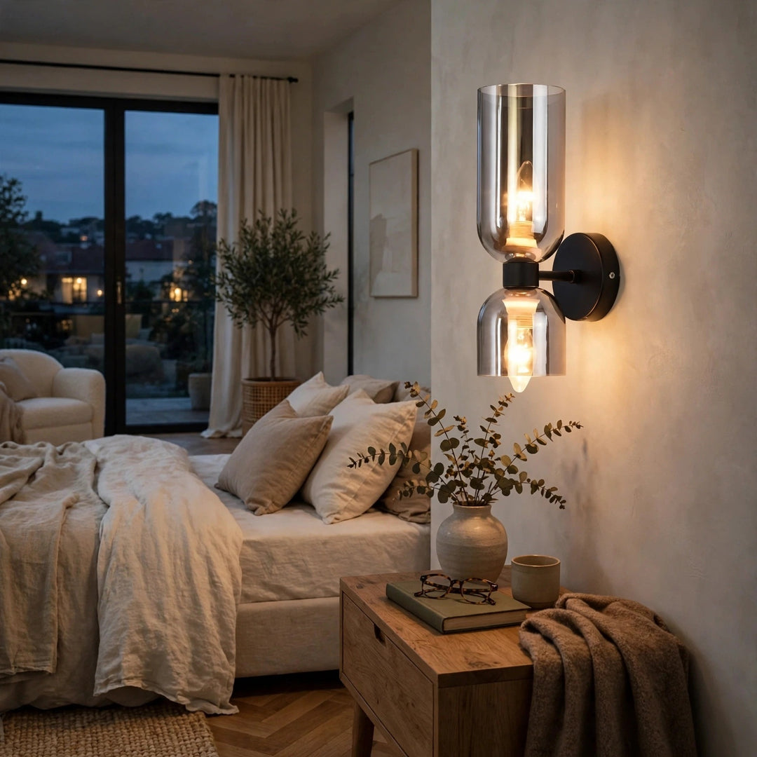 A modern wall-mounted sconce with two clear glass shades and warm exposed bulbs, mounted on a beige wall in a softly lit bedroom.