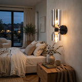 A modern wall-mounted sconce with two clear glass shades and warm exposed bulbs, mounted on a beige wall in a softly lit bedroom.