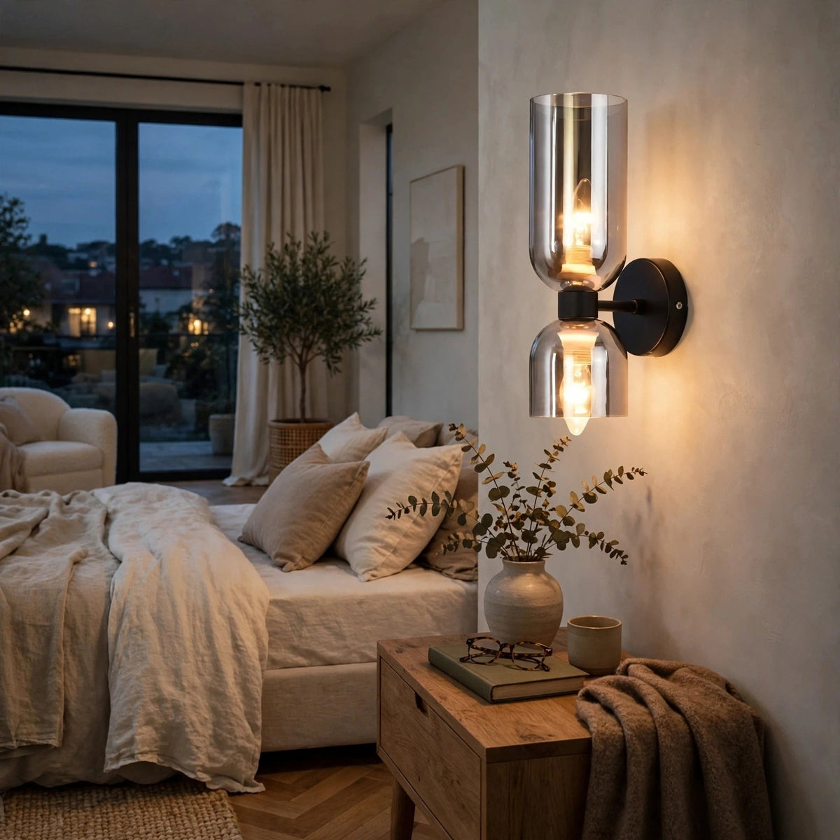A modern wall-mounted sconce with two clear glass shades and warm exposed bulbs, mounted on a beige wall in a softly lit bedroom.