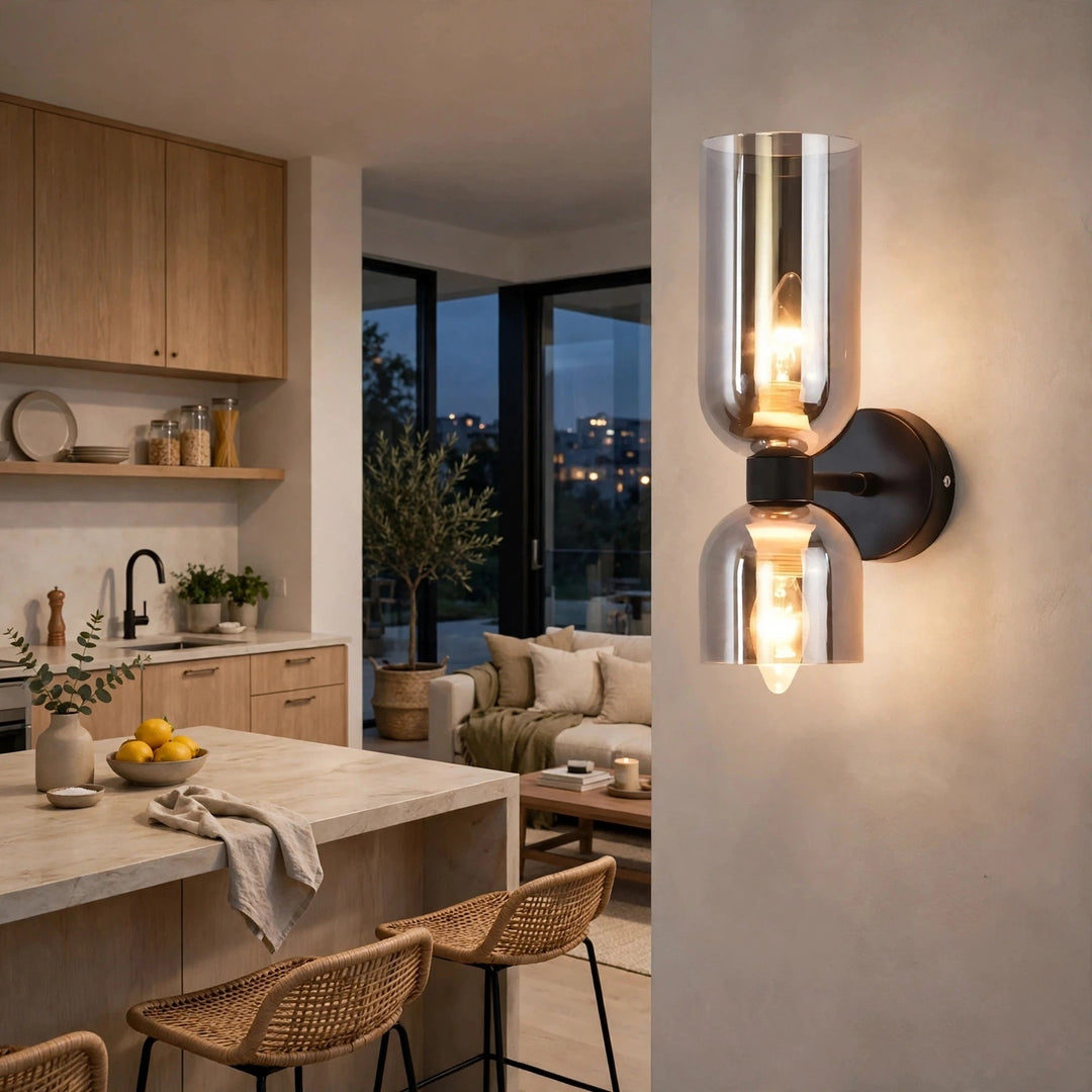 A modern wall-mounted sconce light fixture with two clear glass cylindrical shades and glowing Edison-style bulbs, mounted on a beige wall in a contemporary kitchen and living room setting.