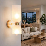A modern wall-mounted sconce with two frosted cylindrical shades and a brushed gold metal base, shown lit in a contemporary living room setting.
