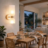 A modern wall-mounted sconce with two glowing cylindrical frosted shades and a brushed gold metal base, installed on a textured off-white wall in a beautifully styled dining room.
