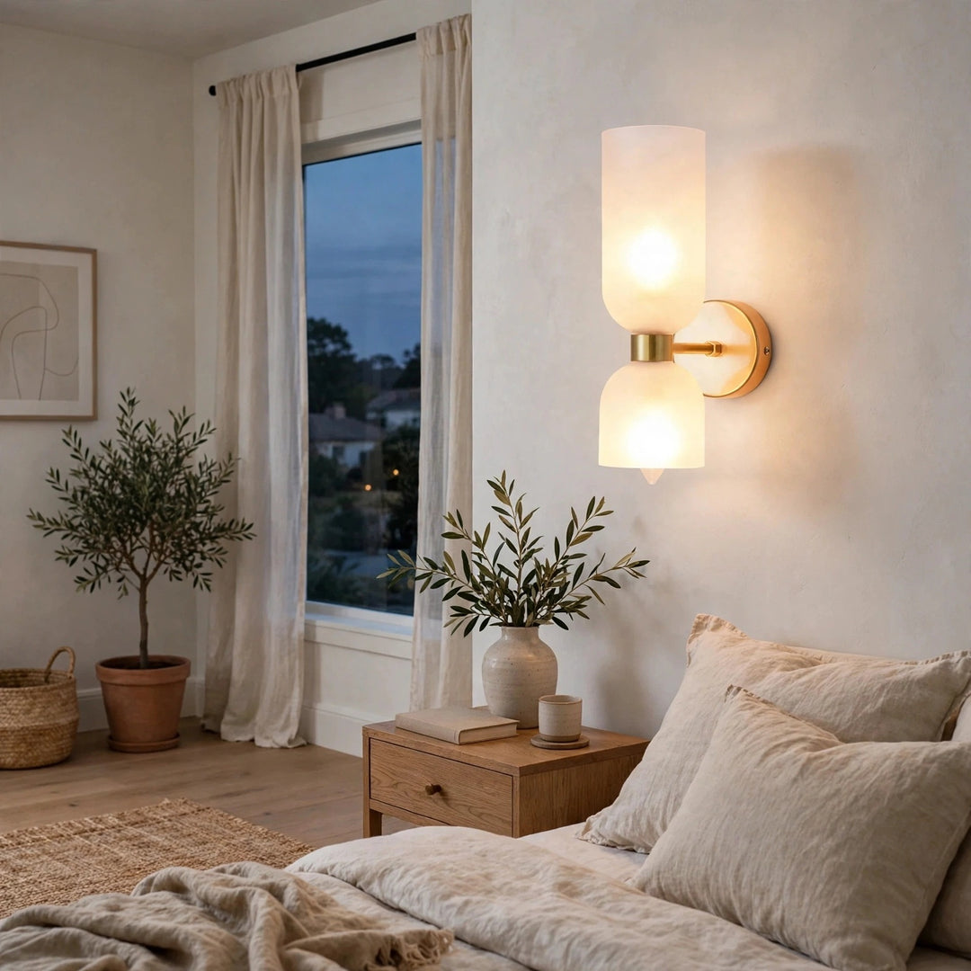 A modern wall-mounted sconce with two frosted cylindrical shades and a brushed brass center cap, glowing warmly in a softly lit bedroom setting.
