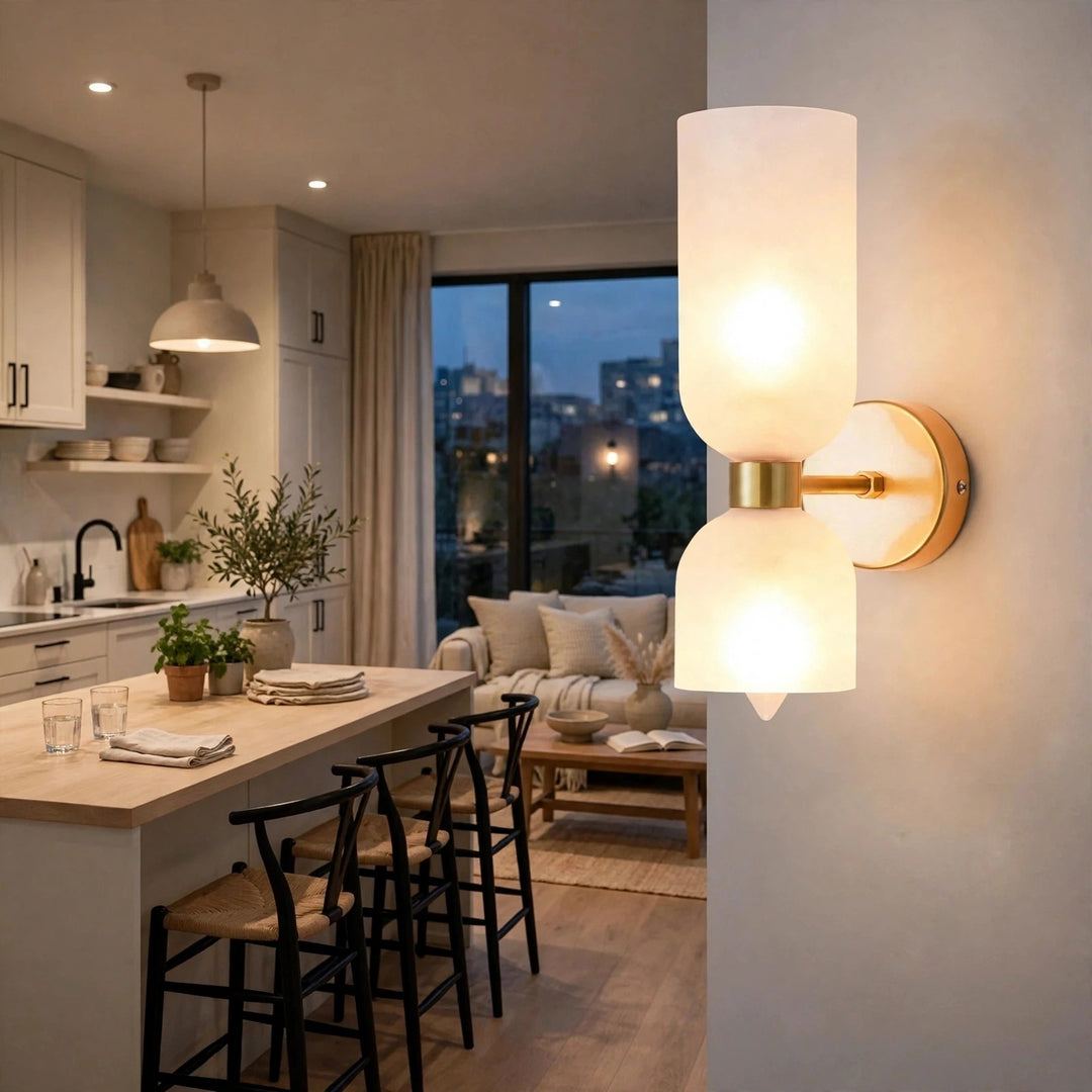 Gold-tone wall sconce with two glowing frosted white glass shades mounted beside an open-plan kitchen and living area.