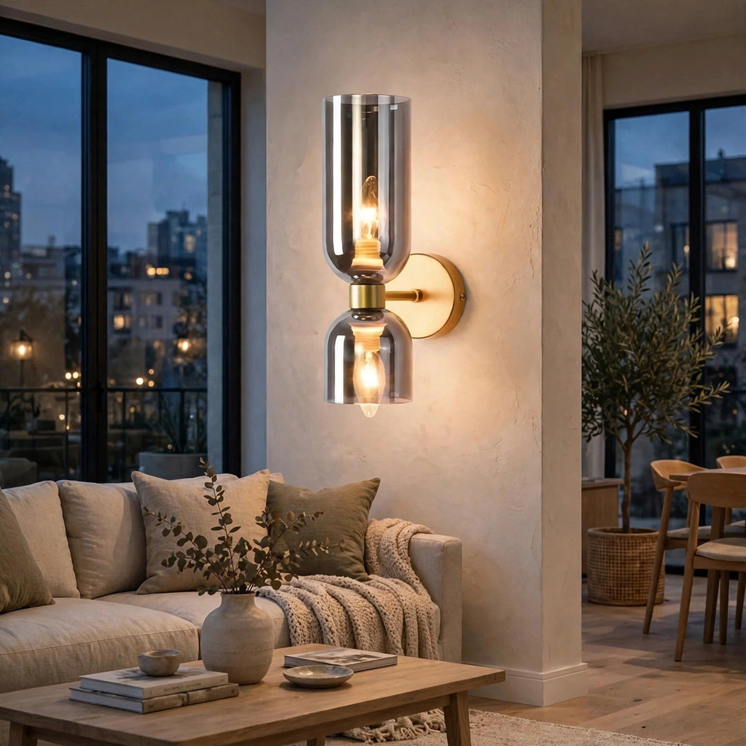 A modern wall-mounted sconce with two clear glass shades and warm Edison bulbs, mounted on a beige wall in a stylishly decorated living room.