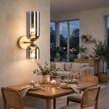 A modern wall-mounted sconce with two vertical brushed-metal shades and glowing exposed bulbs, installed on a warm beige wall in a styled dining and living room setting.