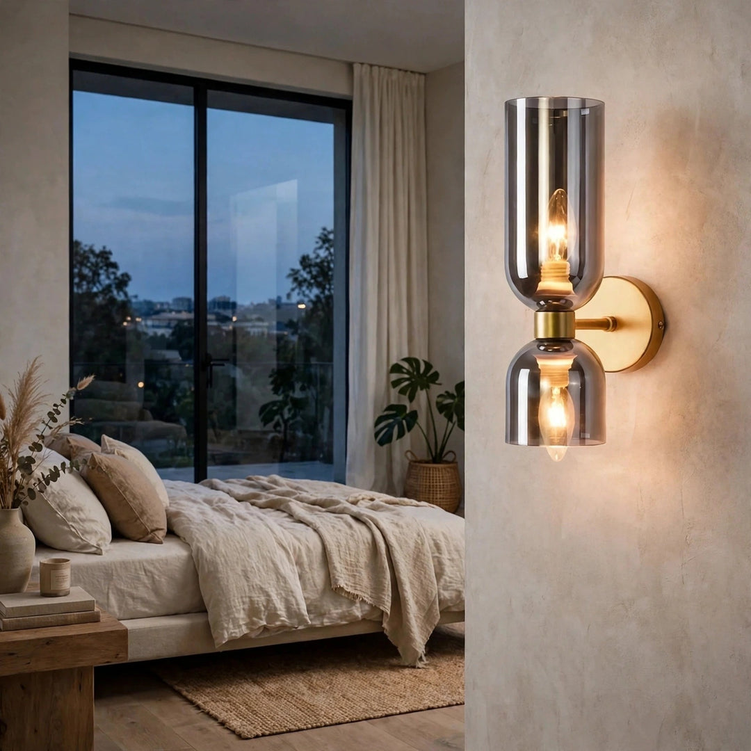 A modern double-light wall sconce with a brushed silver chrome finish and warm glowing bulbs, mounted on a textured beige wall in a styled bedroom setting.