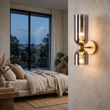 A modern double-light wall sconce with a brushed silver chrome finish and warm glowing bulbs, mounted on a textured beige wall in a styled bedroom setting.