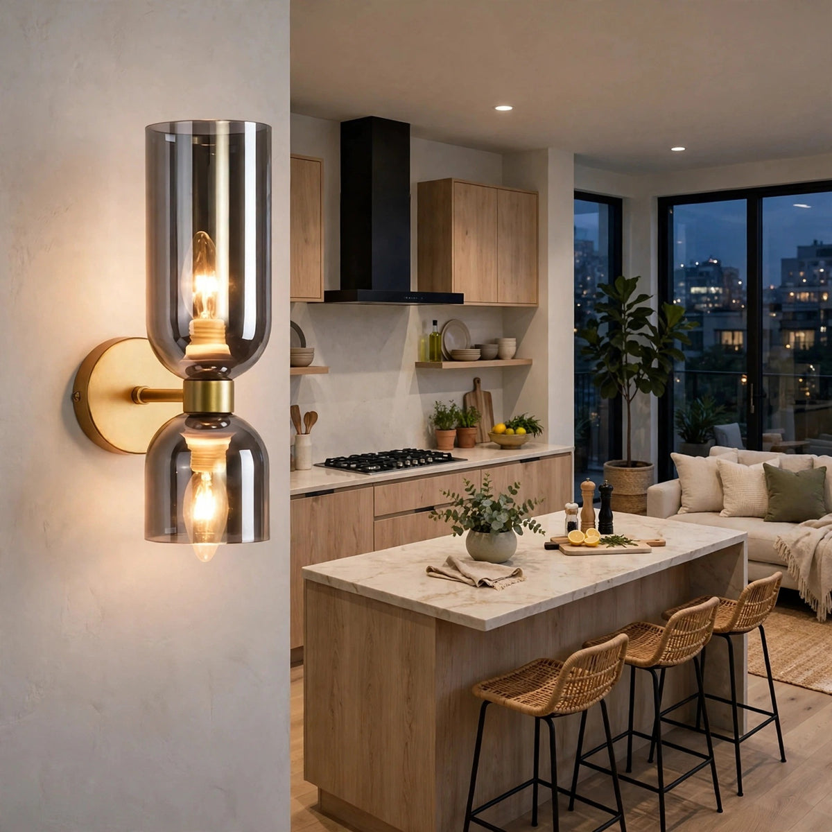 A modern wall-mounted sconce with two clear cylindrical glass shades and warm Edison-style bulbs, shown lit in a real kitchen interior with wood cabinetry, island, stools, and large windows.