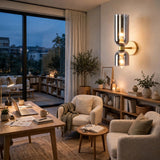 A modern wall-mounted sconce with two stacked cylindrical glass shades and glowing warm bulbs, mounted on a beige wall in a cozy, warmly lit living room with armchairs, a wooden desk, and plants.