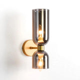 A modern wall-mounted sconce light fixture with two smoked glass shades and a brushed brass backplate and central collar, shown against a plain white wall.