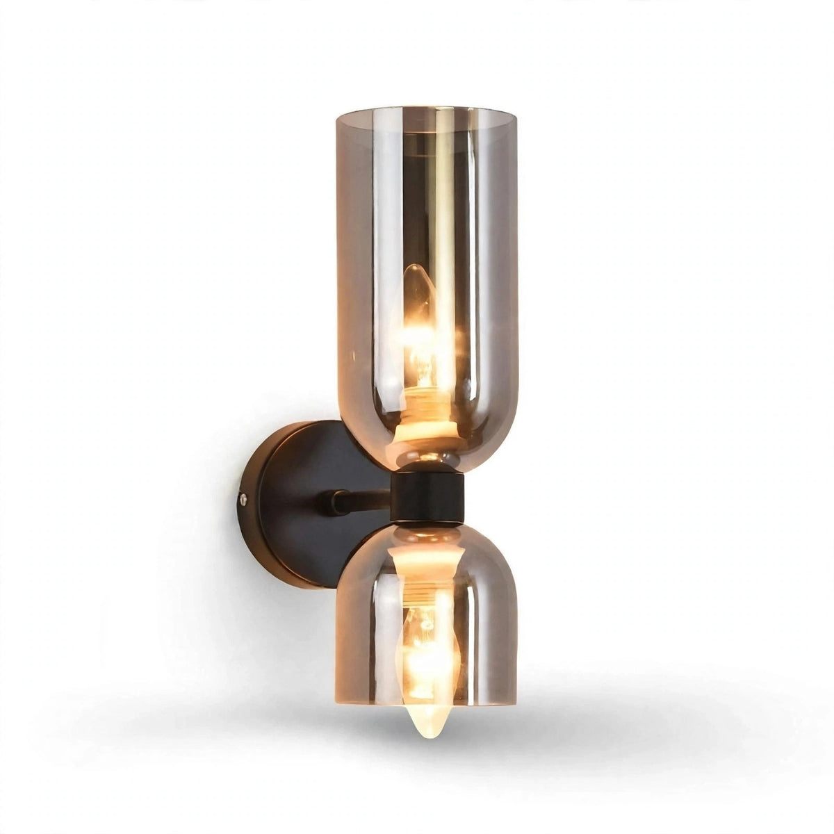 A modern industrial-style wall sconce featuring two cylindrical glass shades in a smoky bronze finish, with a warm glowing light bulb visible inside, mounted on a dark metal wall plate against a white background.