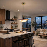 A modern kitchen island with a white marble countertop and dark wood cabinetry, featuring black bar stools under a brass and black pendant light. The background shows a bright, contemporary living room with large windows overlooking a city skyline at dusk.